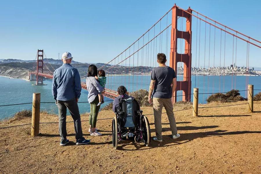 Eine Gruppe von Menschen, 包括一个坐轮椅的人, ist von hinten zu sehen, 从马林岬俯瞰金门大桥。.