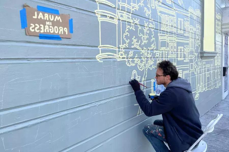 一名艺术家在Mission District一幢建筑物的一侧画壁画, 与一个标志在建筑上说“壁画正在进行中”. San Francisco, CA.