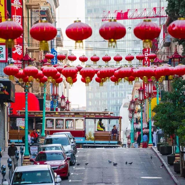 Uma rua montanhosa na 唐人街 de São Francisco é retratada 和 lanternas vermelhas penduradas e um bonde passando.